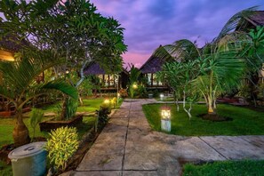 Un sendero conduce a un resort con estructuras de techo de paja, rodeado de exuberante vegetación y céspedes bien cuidados.