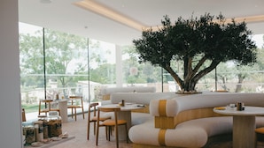 A modern restaurant with a large tree centerpiece, cushioned seating, and a buffet area with food containers. - La Maison Verchant (Montpellier)