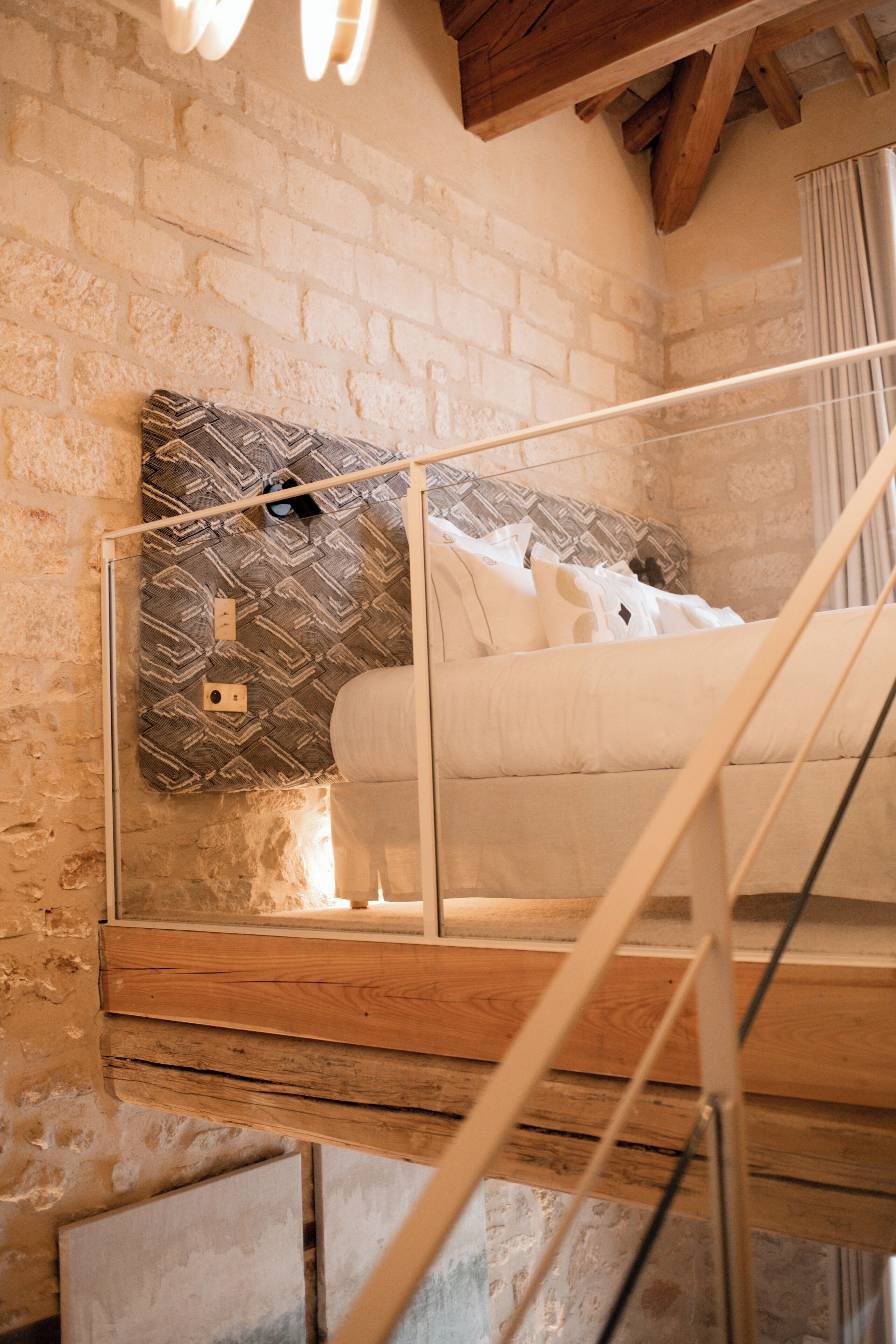 A loft bedroom with exposed brick walls, a wooden ceiling, and a glass railing.