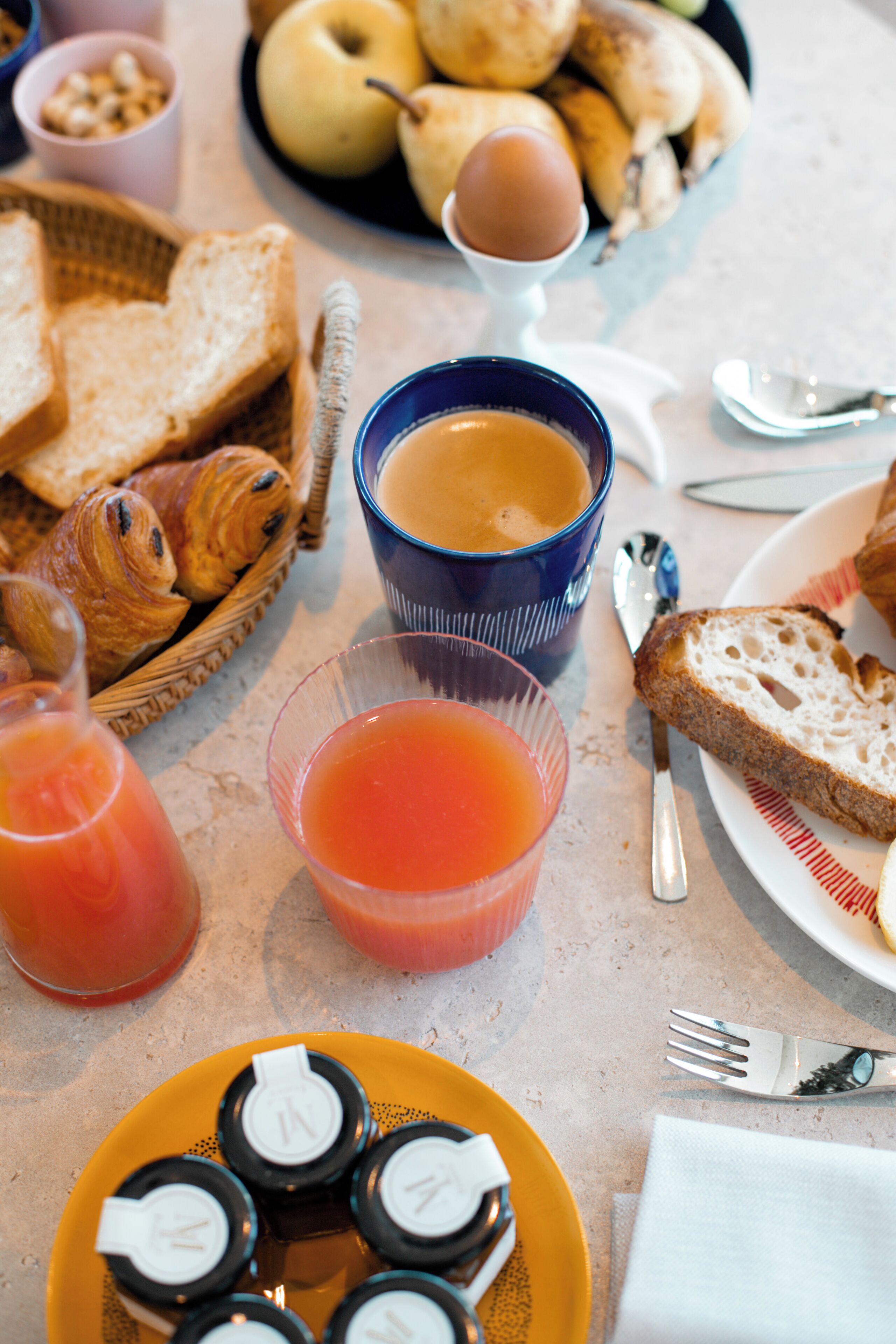 Una mesa de desayuno con cruasanes, pan, un plato de fruta, una taza de café y un vaso de jugo de naranja.
