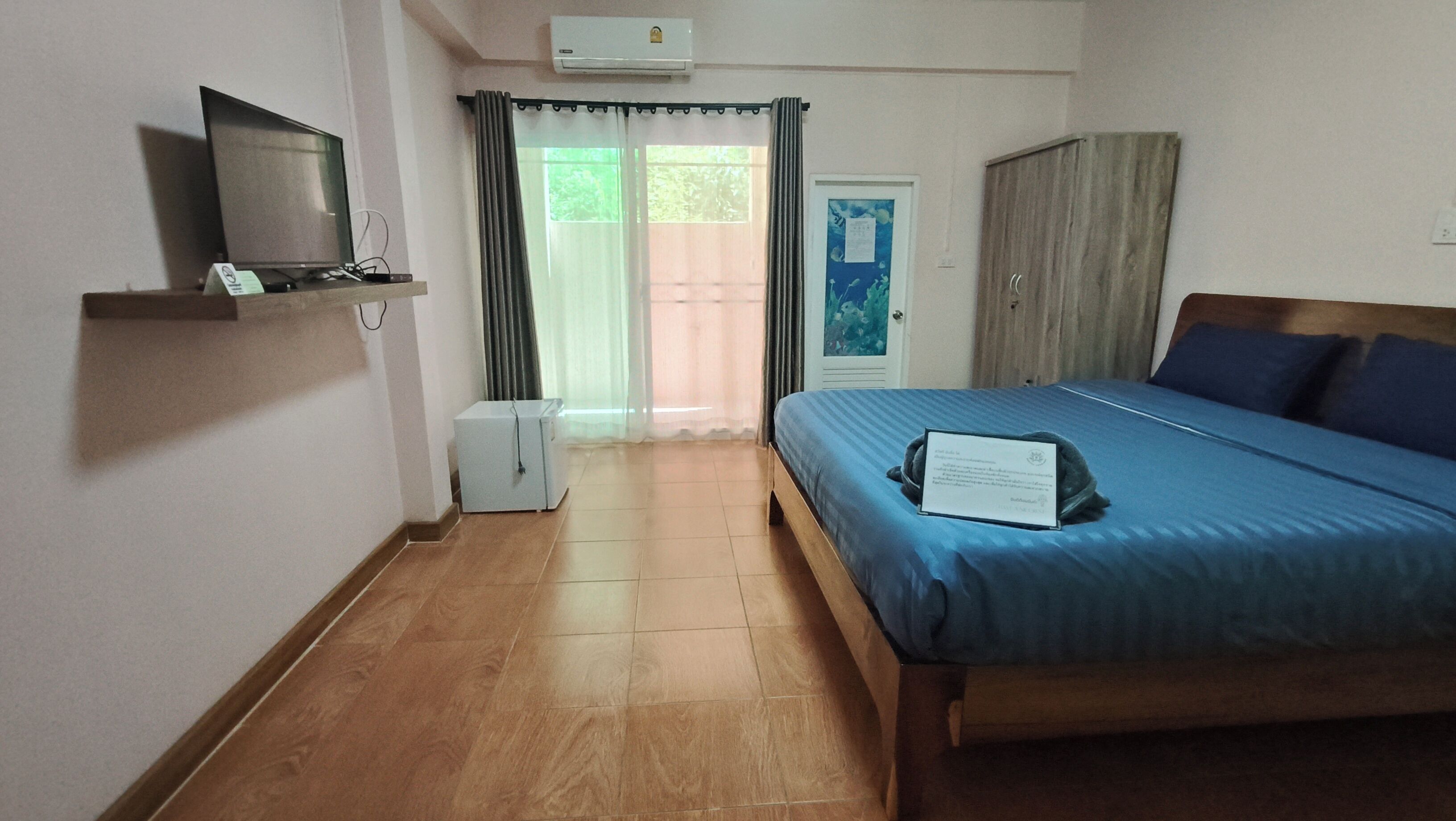 A bedroom with a bed, a television on a shelf, a small refrigerator, and a door leading to a balcony.