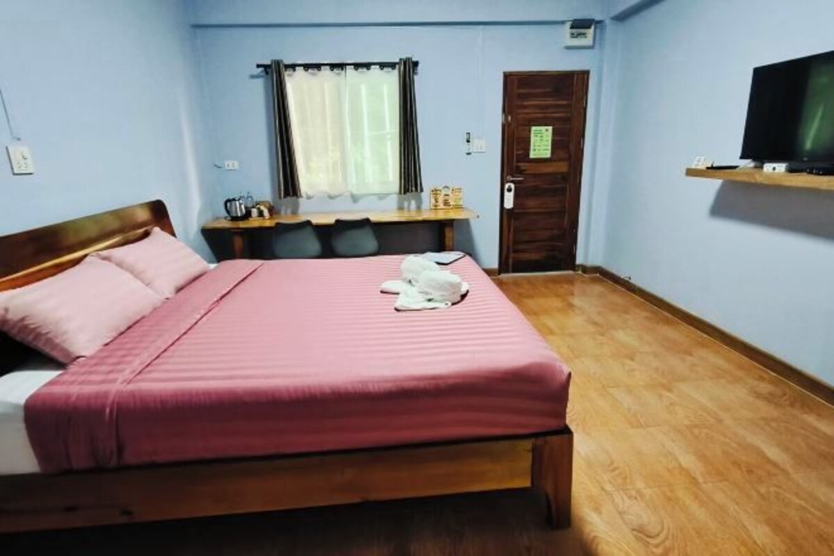 A bedroom with a wooden bed, a desk with a chair, a television on a shelf, and a window with curtains.
