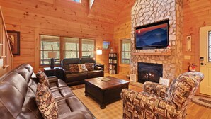 A cozy log cabin interior with a stone fireplace, leather furniture, and a television mounted on the wall.
