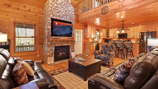 A cozy cabin interior with a stone fireplace, leather sofas, a dining area, and a kitchen.