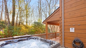 A hot tub on a wooden deck with a view of trees and a wooden building in the background.