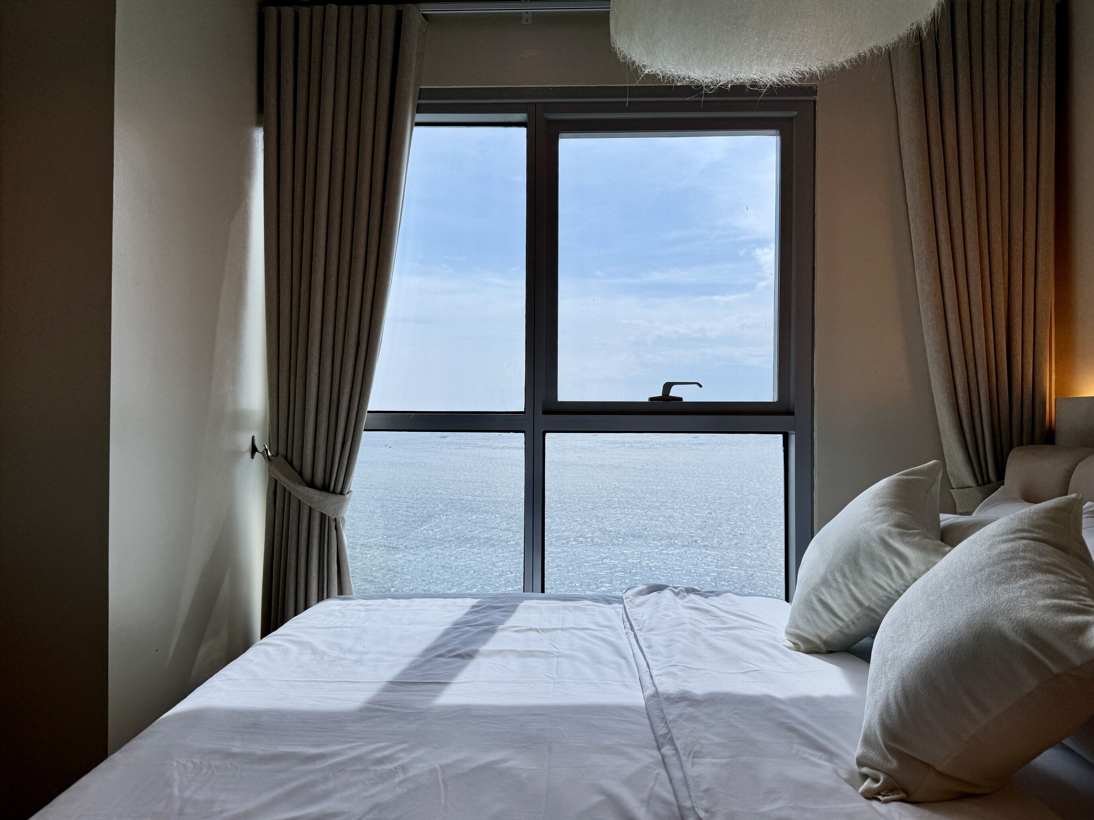 A hotel room with a large window, a bed with white linens, and a view of the sea.