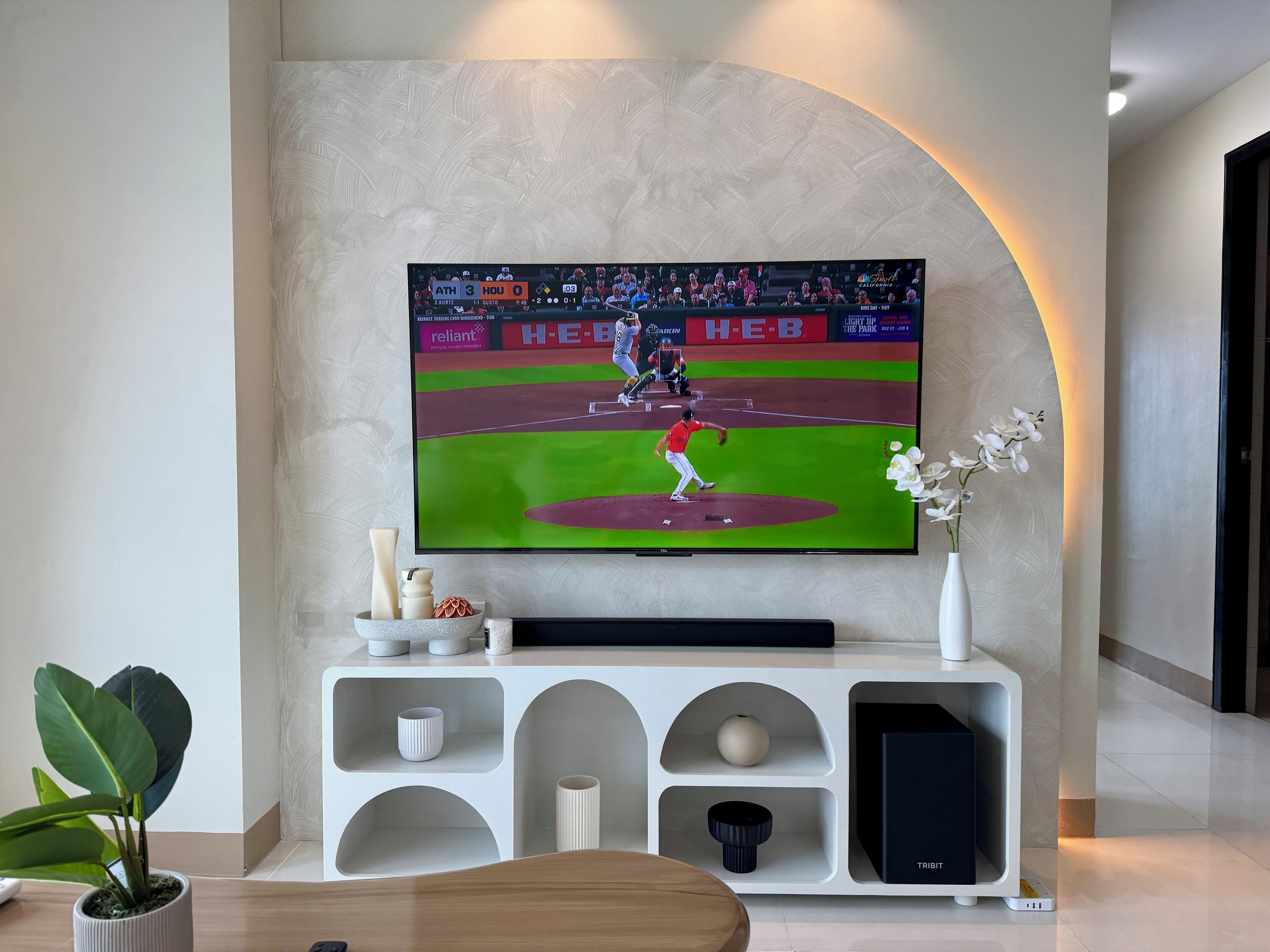 A flat-screen TV mounted on a wall displaying a baseball game, with a white console below featuring decorative items and a vase with flowers.