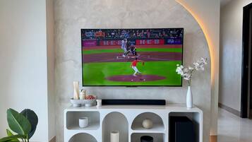 A flat-screen TV mounted on a wall displaying a baseball game, with a white console below featuring decorative items and a vase with flowers.