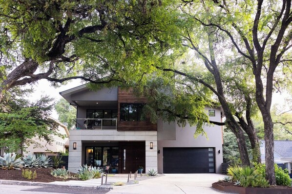 A modern two-story house with a garage, surrounded by mature trees and landscaped garden beds.