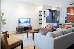 A modern living room with a flat-screen TV, a sofa, a coffee table, and a dining area with a wooden accent wall. - *NEW Central Austin | Sleeps 12 - WalkerVR Firefly (Austin)