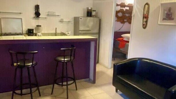 A compact kitchen with a bar area, two stools, a black sofa, and a ceiling fan. - Air conditioned home w/ full kitchen, TV & parking (Puerto Viejo de Talamanca)