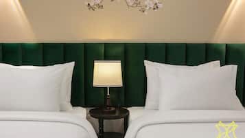 Una habitación de hotel con un cabecero tapizado en verde, dos camas blancas con almohadas a juego, una mesita de noche redonda con lámpara y un adorno floral en la pared.