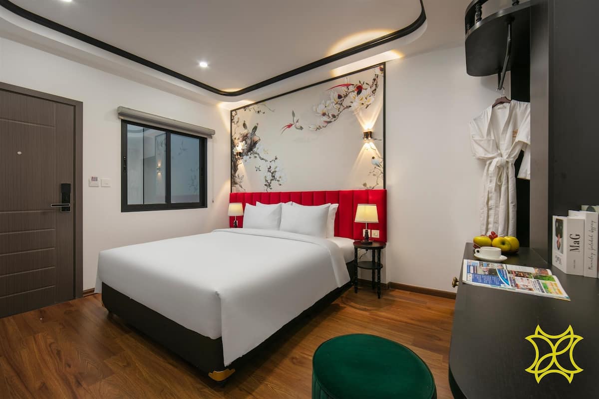 A hotel room with a large bed, a red headboard, a wall-mounted TV, a small table with a lamp, and a green ottoman.