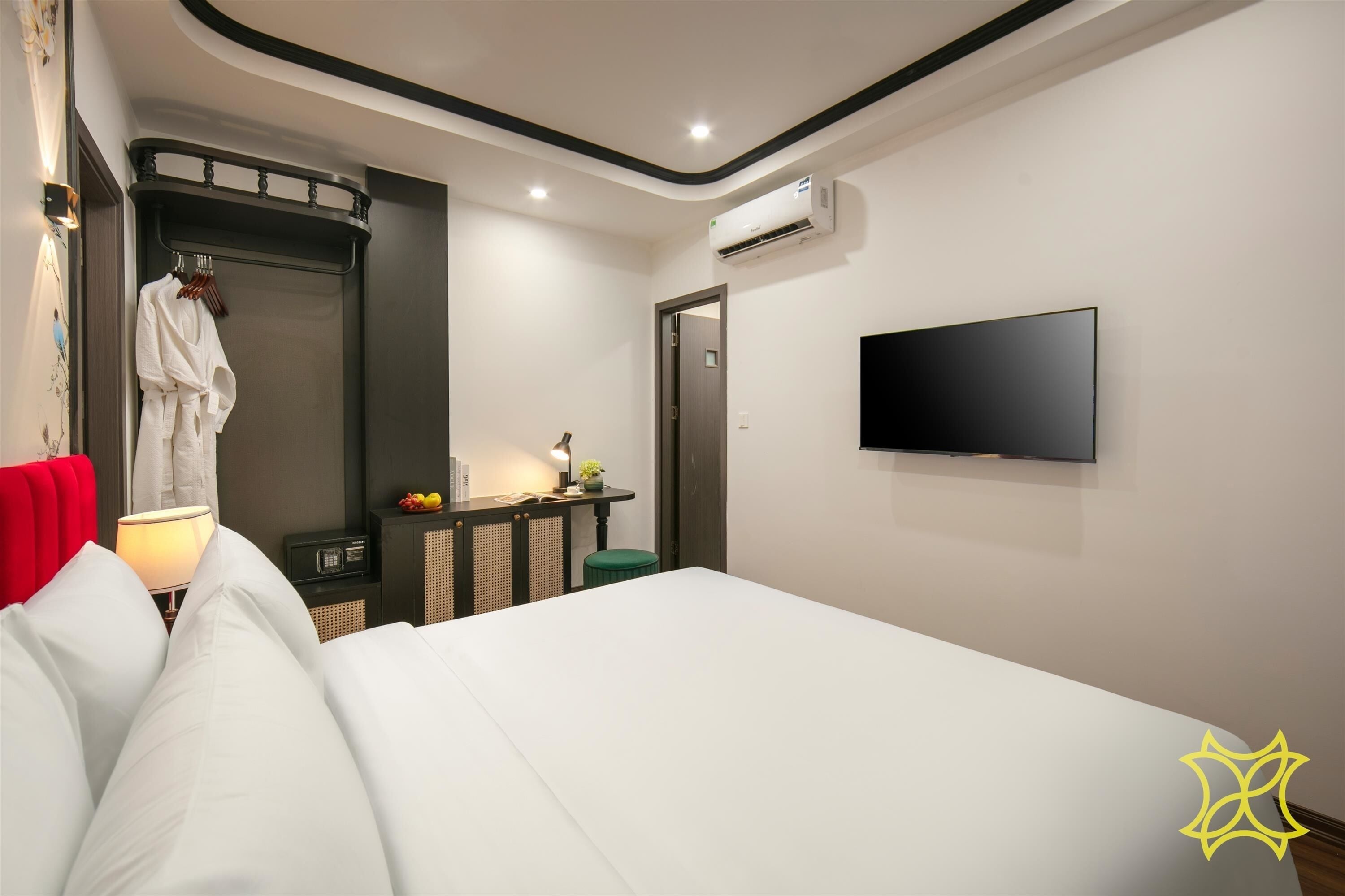 A modern hotel room with a large bed, a flat-screen TV mounted on the wall, a bedside table with a lamp, a small green stool, and a robe hanging on a hook.