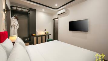 A modern hotel room with a large bed, a flat-screen TV mounted on the wall, a bedside table with a lamp, a small green stool, and a robe hanging on a hook.