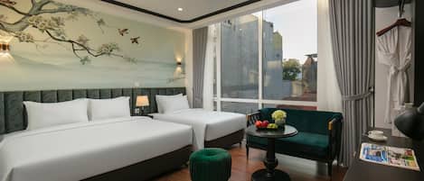 A hotel room with two beds, a green ottoman, a small table with fruit, a ceiling fan, and a large window with curtains.
