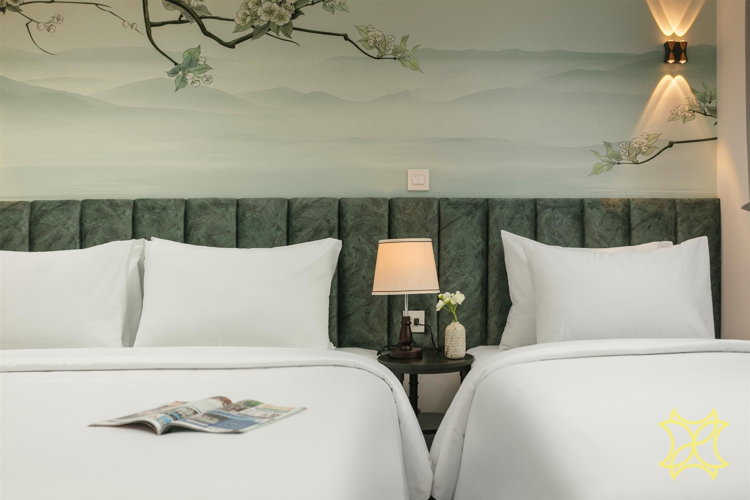 A hotel room with two beds, a nightstand with a lamp, a vase with flowers, and a wall mural.