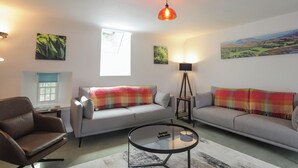 A living room with two grey sofas, a brown armchair, a glass coffee table, and a floor lamp. There are three paintings on the walls and a window with blinds. - Srondubh (Achnasheen)