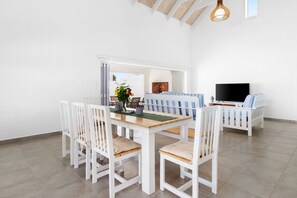 Un comedor con mesa de madera y sillas blancas, un sofá con cojines a rayas y un televisor sobre un mueble.