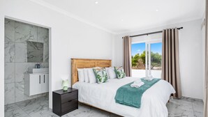 A bedroom with a large bed, a nightstand, a wooden headboard, a bathroom with marble tiles, and a view of greenery through the window. - Sunlit Oasis Ocean and Golf Retreat (St. Helena Bay)