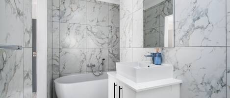A modern bathroom with marble tiles, a freestanding bathtub, and a white vanity with a mirror.