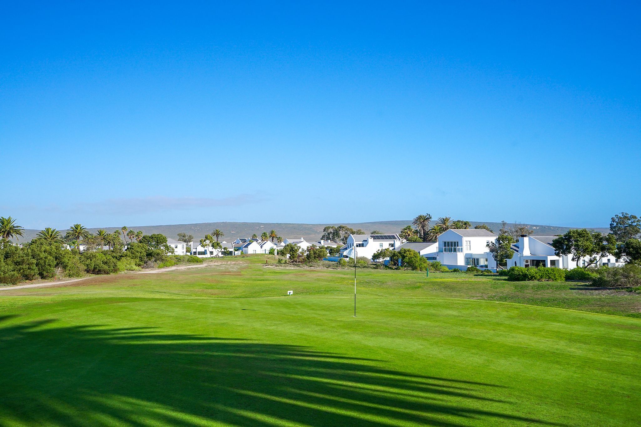 A golf course with a flagstick, surrounded by white houses, palm trees, and a clear blue sky.