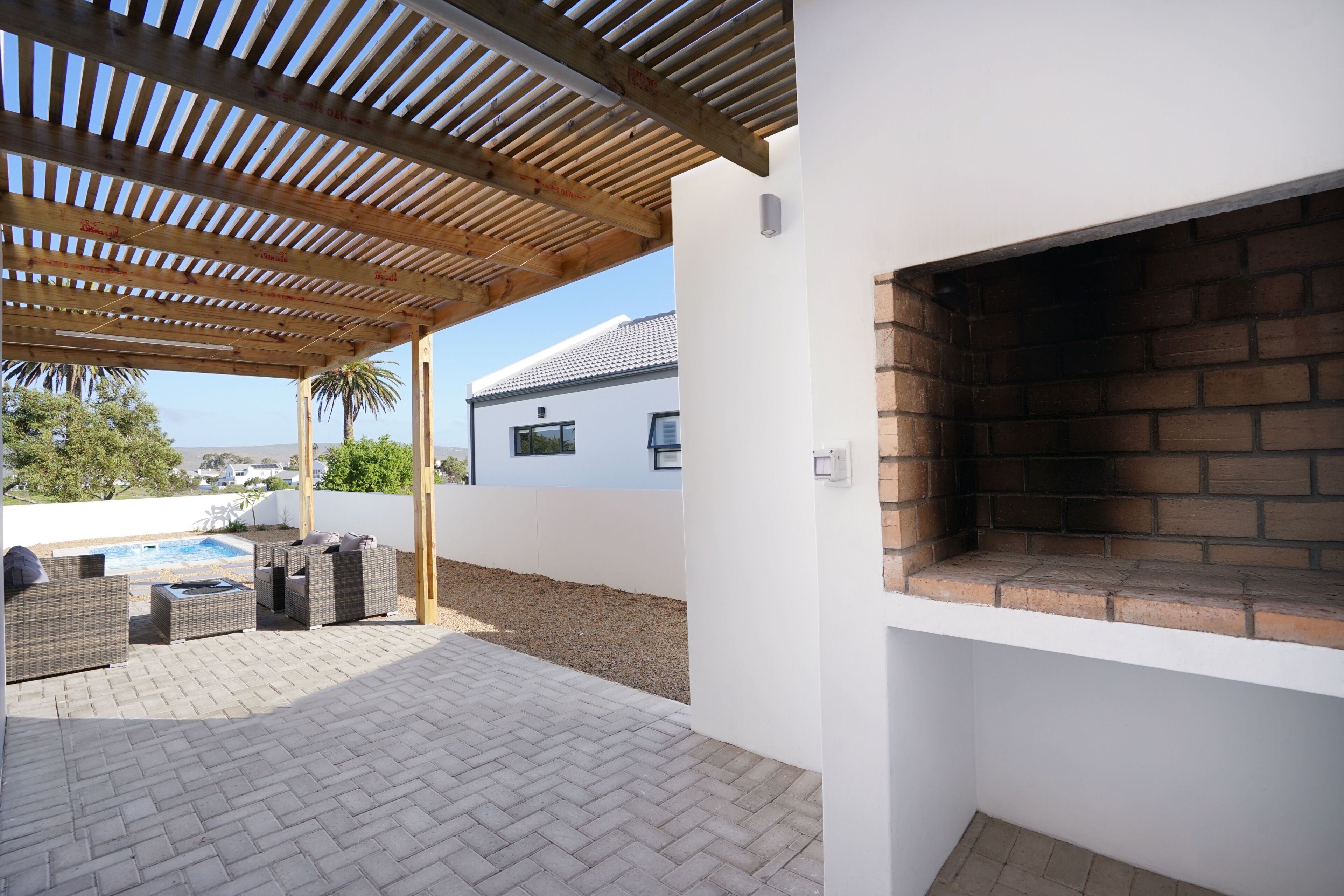 Une terrasse extérieure couverte, dotée d’une cheminée en briques, de meubles en osier et d’une piscine en arrière-plan.