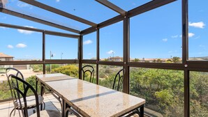 Quarto iluminado pelo sol com uma mesa e cadeiras, grandes janelas revelam vista para vegetação e edifícios ao longe.