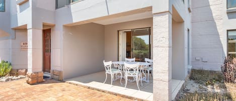 A modern building with a patio area featuring a table and chairs, a glass door entrance, and a sign indicating the address.