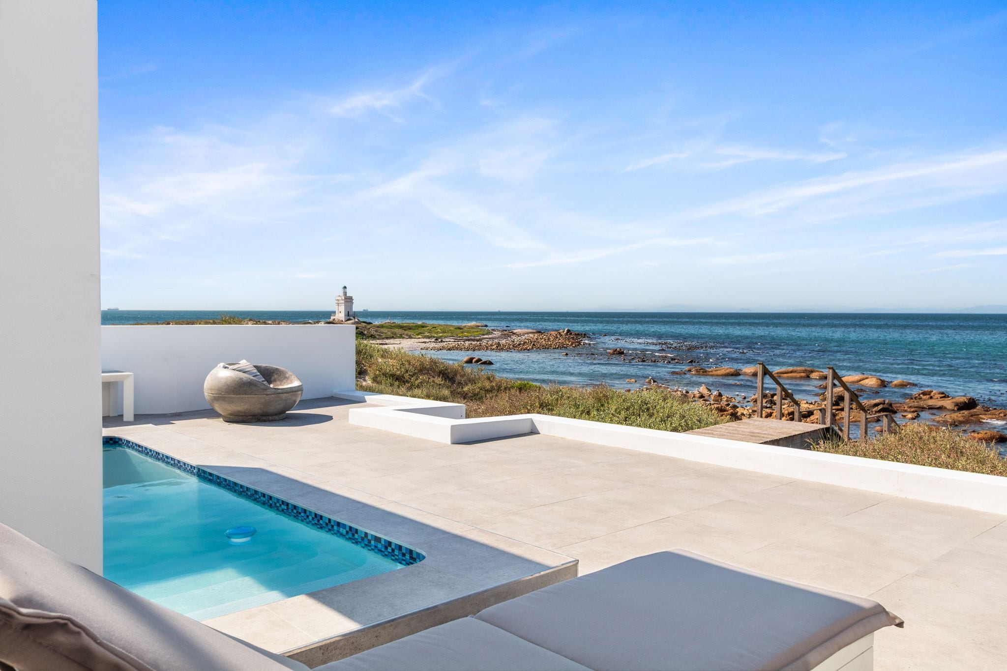 A modern outdoor pool area with a view of the sea and rocky shoreline.