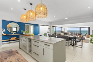 A modern kitchen with a central island, a dining area with a view of the sea, and a living area with a large window overlooking the ocean. - Shelley Point Beach Villa (St. Helena Bay)