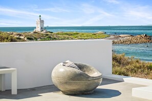 Un espace de détente extérieur moderne, doté d’un fauteuil original en forme de bol en pierre, d’une table blanche et offrant une vue sur la mer et un phare.