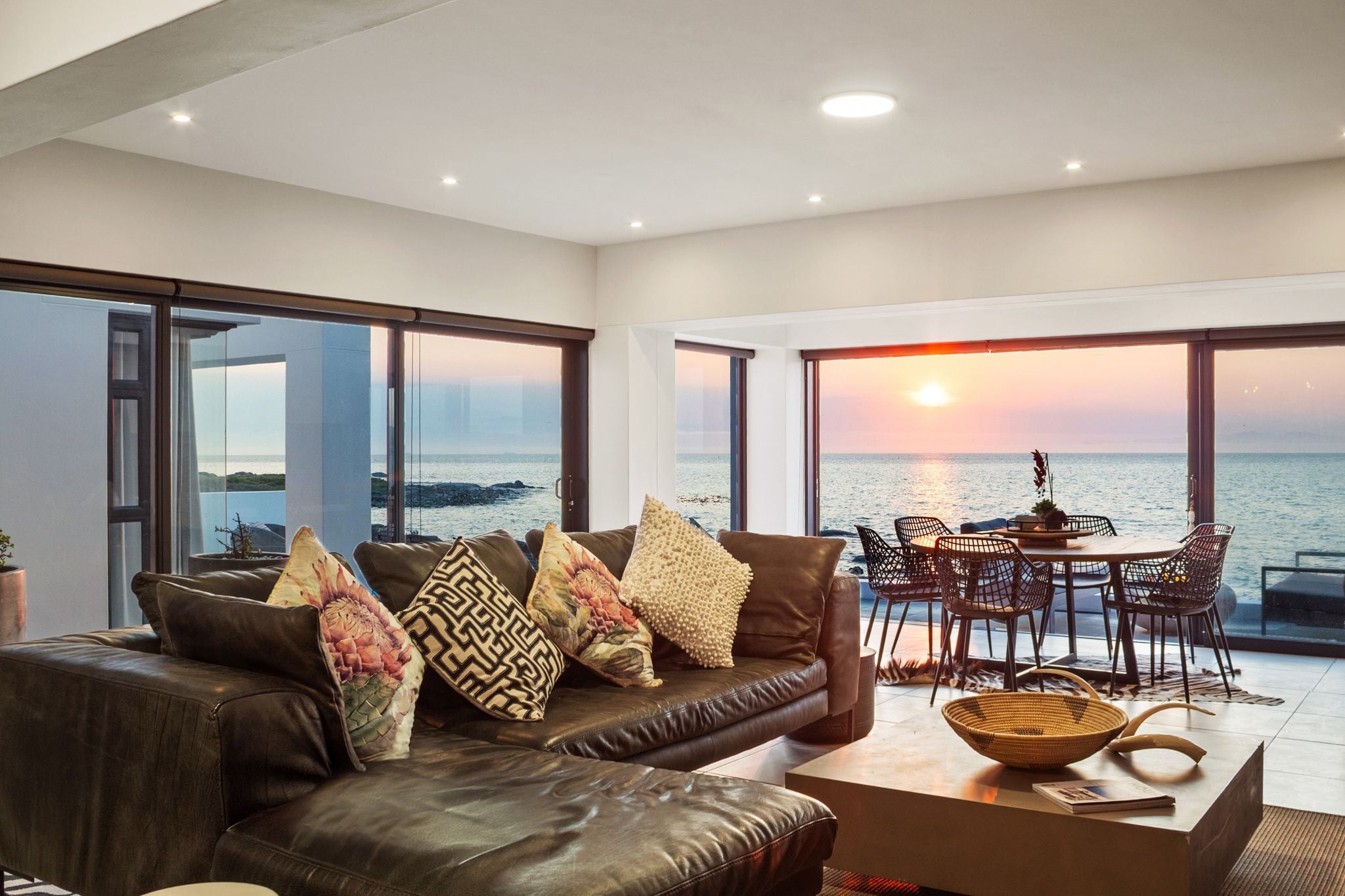 En moderne stue med en lædersofa, et spisebord med stole og store vinduer med udsigt til havet.