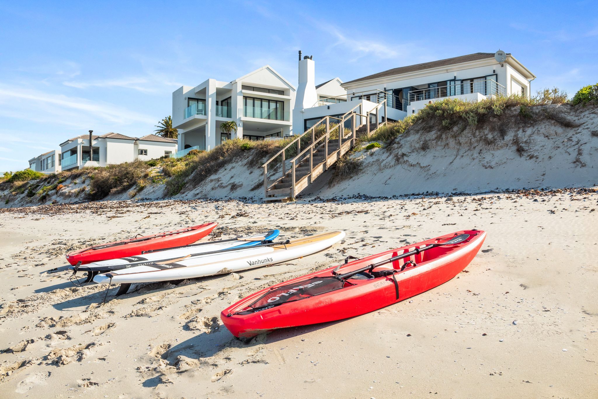 A beachfront property with modern architecture, a staircase leading to a second level, and several kayaks on the sand.