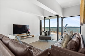 A modern living room with a large flat-screen TV, leather sofas, and a glass door leading to a balcony with a view of the sea. - Shelley Point Beach Villa (St. Helena Bay)