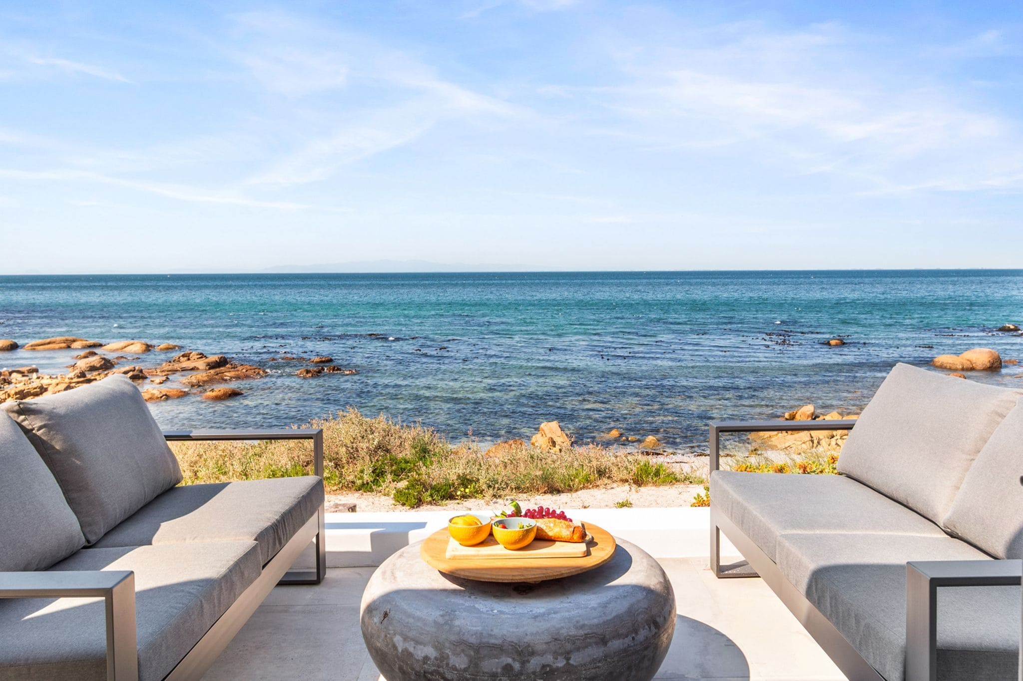 A seaside terrace with two sofas and a table with fruit and a drink, overlooking the sea.