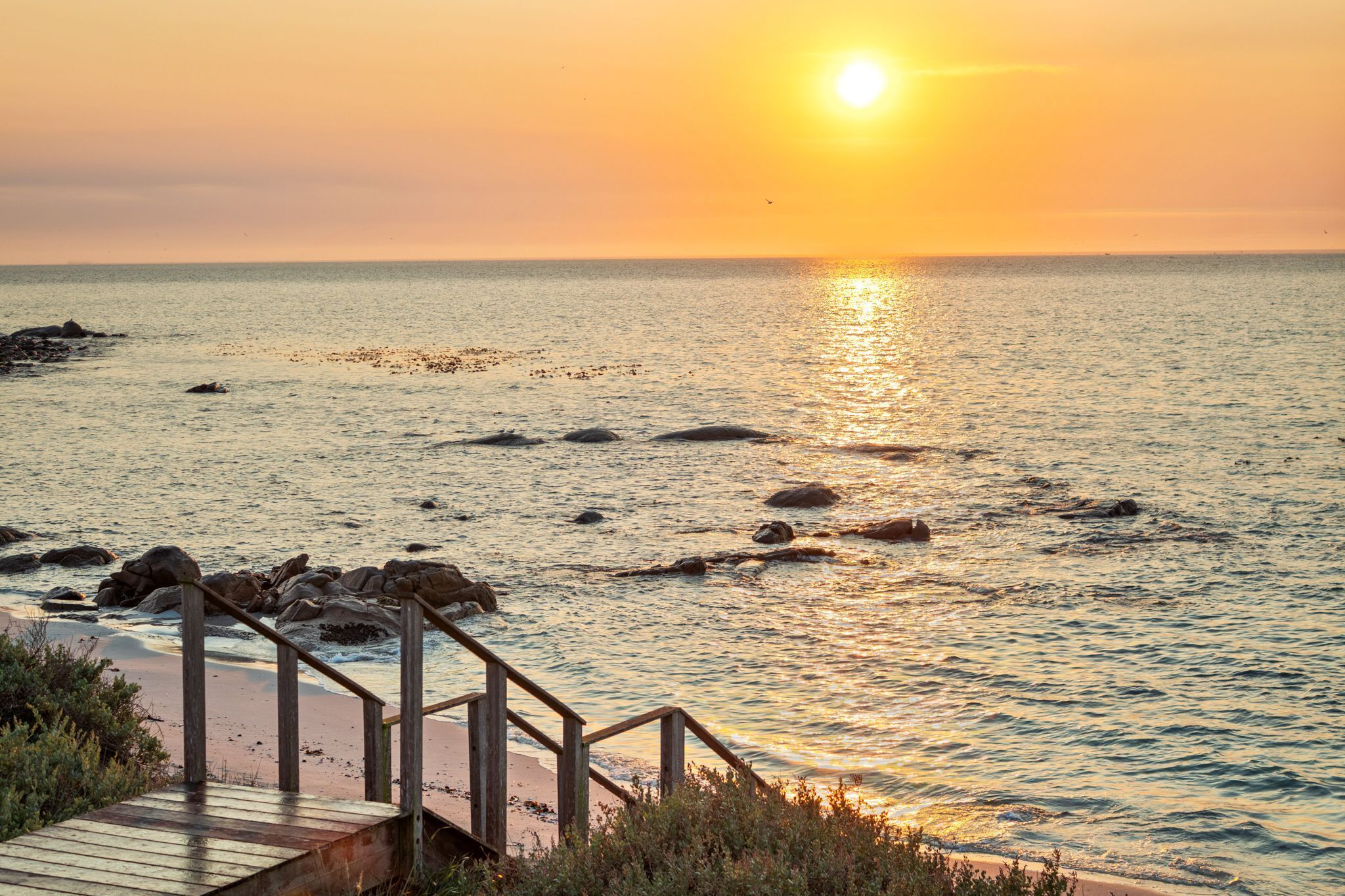 Une terrasse en bois qui mène à une plage, avec un coucher de soleil en arrière-plan.