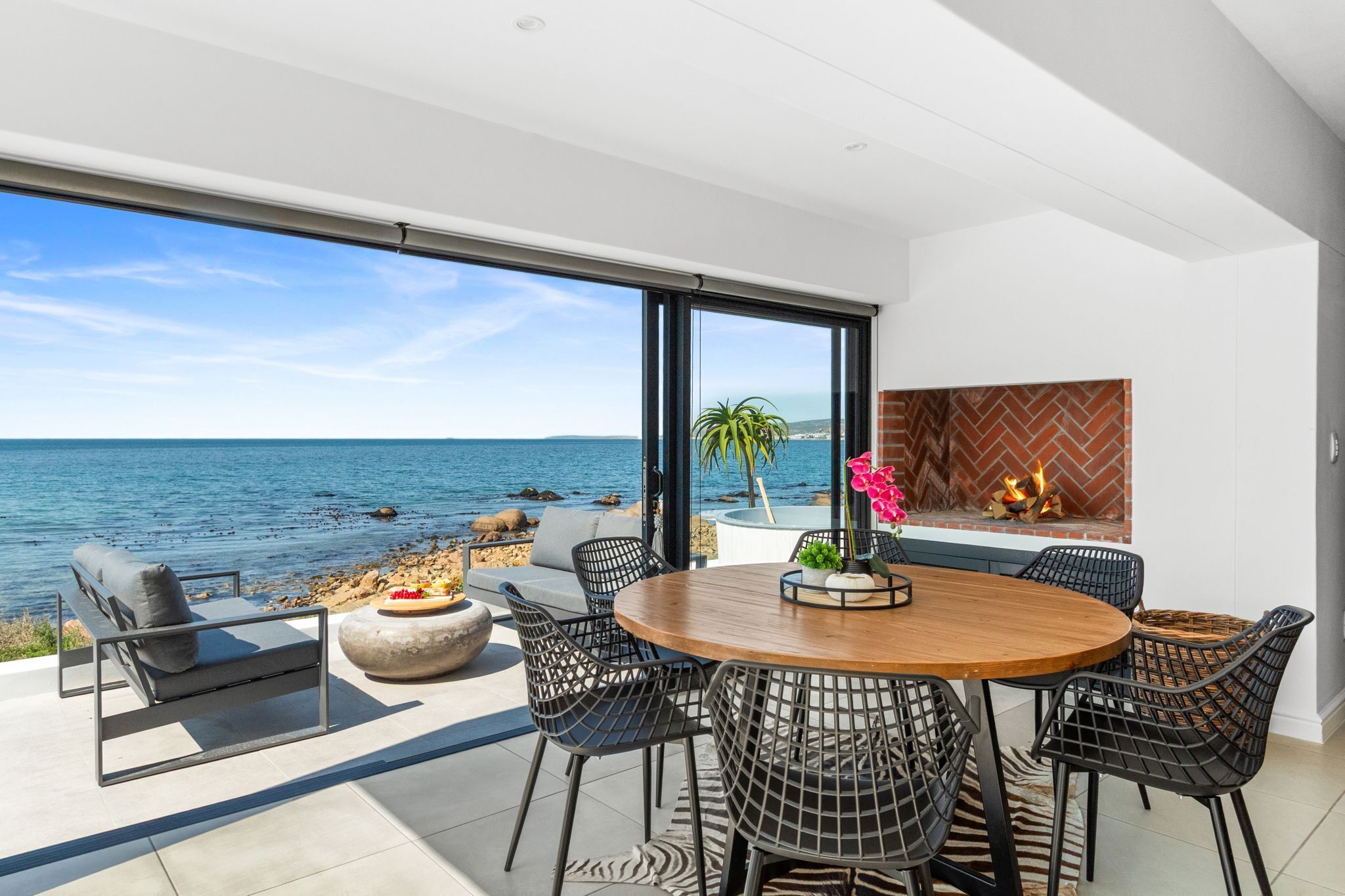 A modern outdoor dining area with a view of the sea, a fireplace, and comfortable seating.
