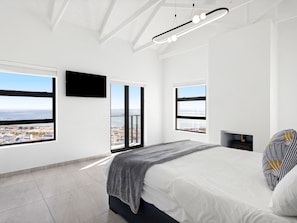 A modern bedroom with a large bed, a TV mounted on the wall, and a view of the city through large windows.