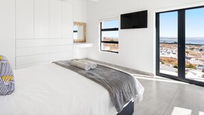 Et moderne soveværelse med en stor seng, udsigt over byen gennem en skydedør og et loft med synlige bjælker.