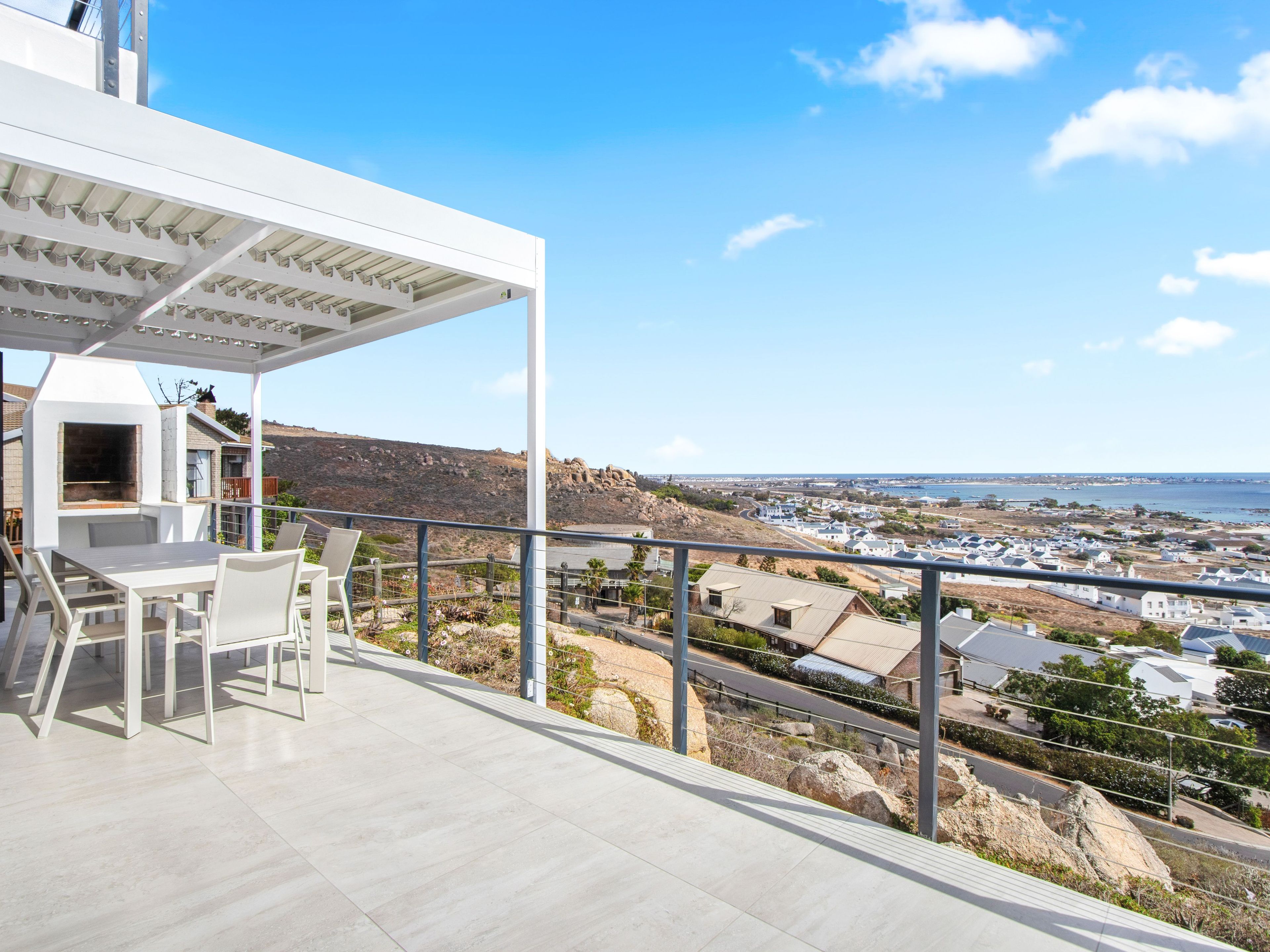 A balcony with a white pergola, outdoor seating, and a view of a coastal town.