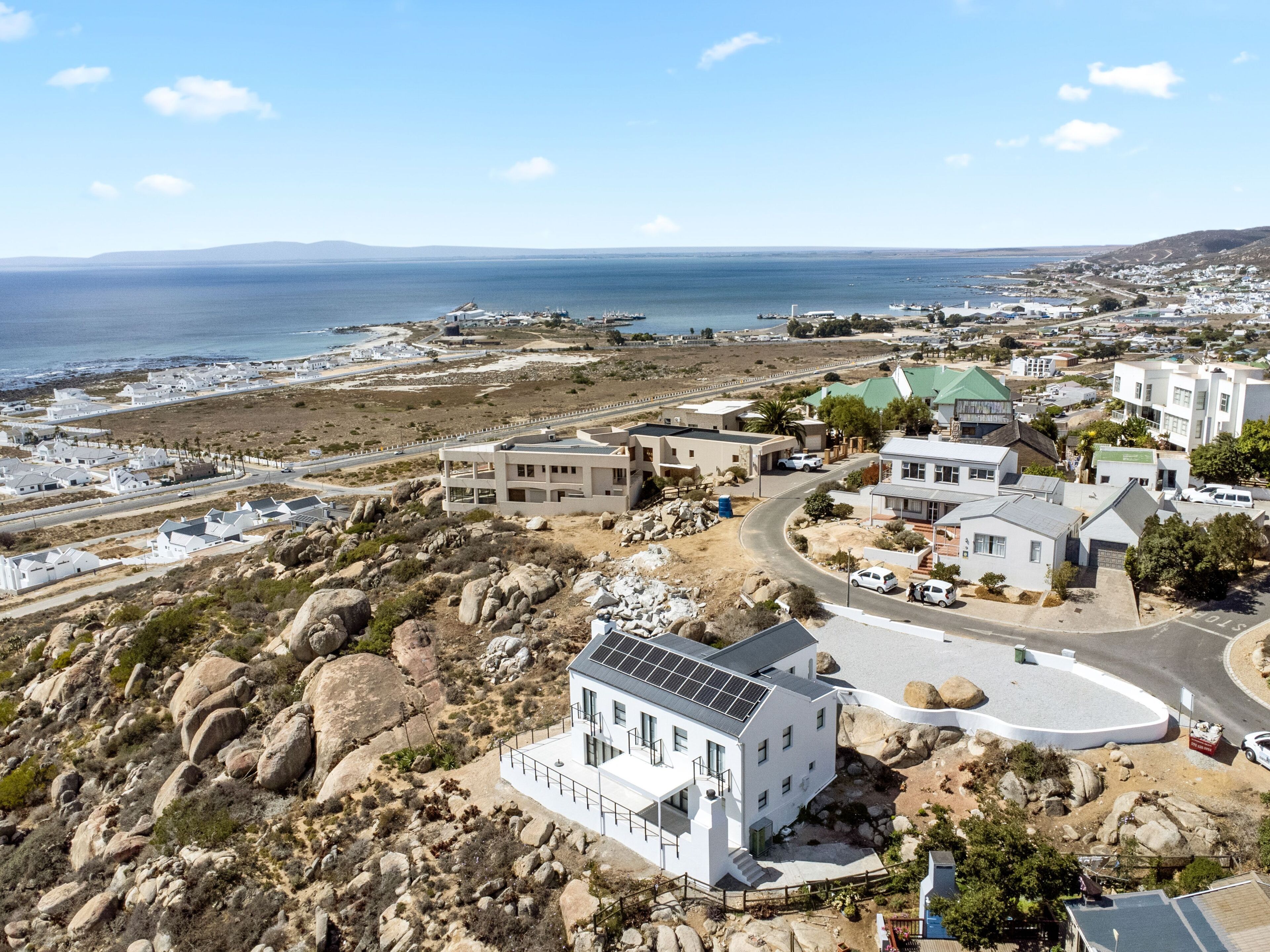 A coastal town with white buildings, a road curving through the center, and a view of the sea and distant hills.