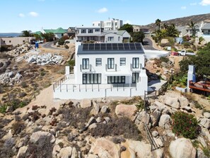 A modern two-story house with a flat roof and solar panels, surrounded by rocky terrain and sparse vegetation.