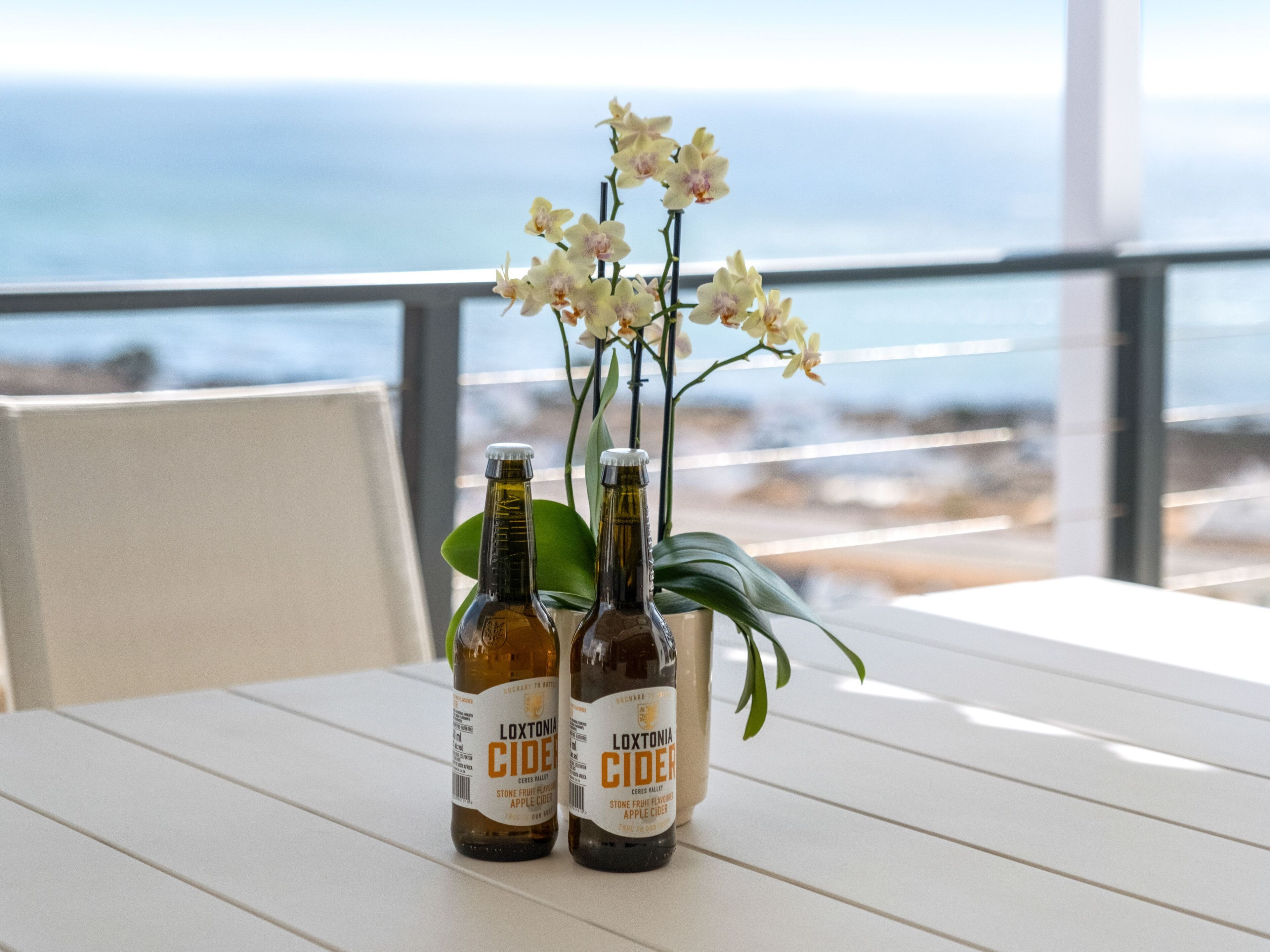 Deux bouteilles de cidre Loxtonia sur une table blanche, avec vue sur l’océan.