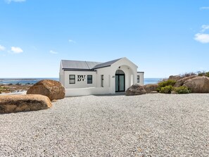A small white building with a black door and windows, labeled 'DV,' situated on a gravel surface with large rocks and a clear view of the sea in the background.