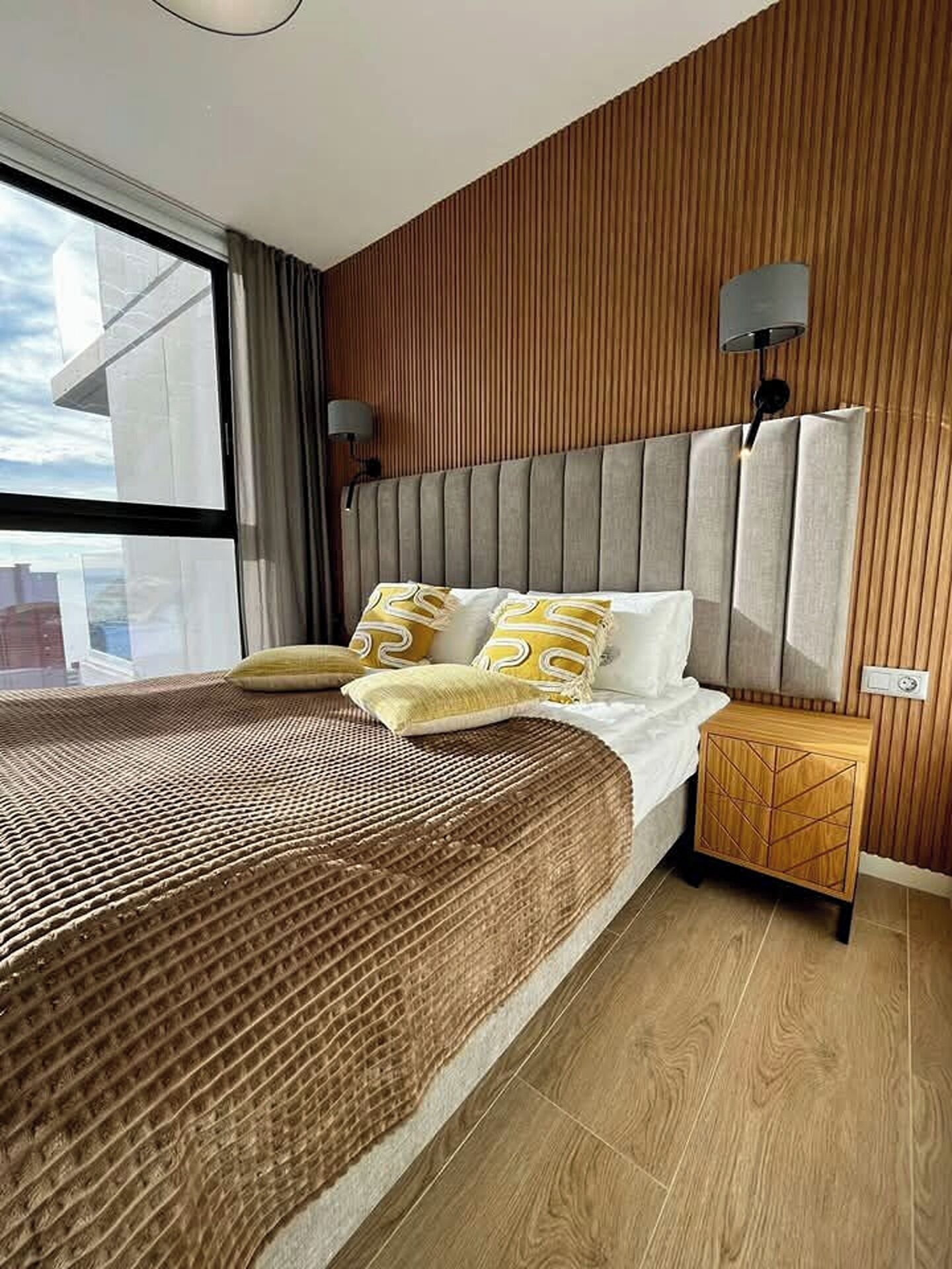 A modern bedroom with a large bed, wooden panel walls, and a nightstand with a lamp.
