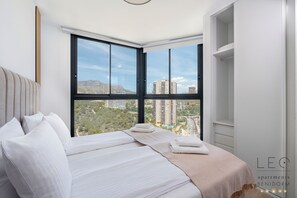 A bedroom with a large window, a bed with white linens, and a view of a cityscape. - Torre Sakura By Leo Apartments (Benidorm)
