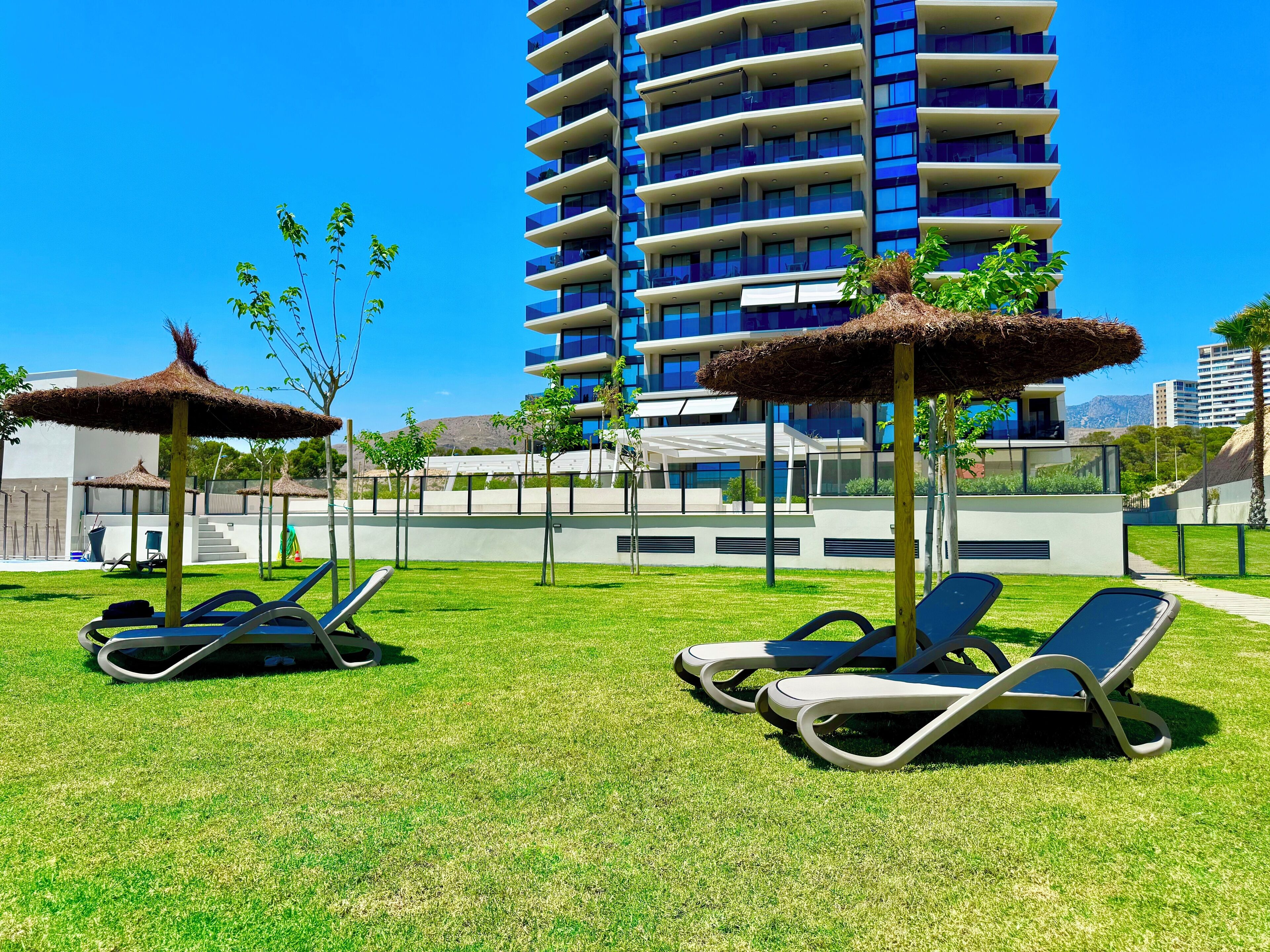 A modern outdoor lounge area with sun loungers and thatched umbrellas, surrounded by a well-maintained lawn and a tall building in the background.