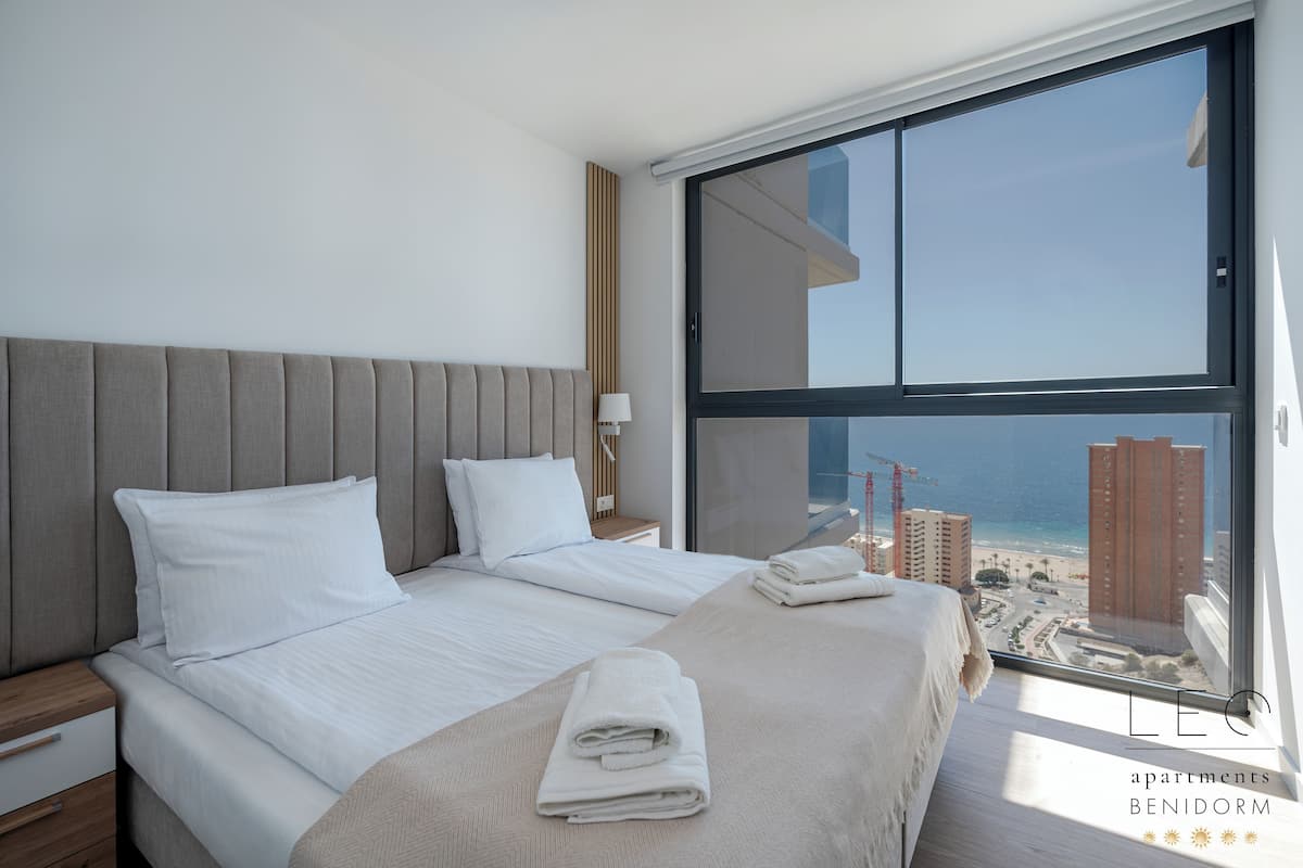 Un dormitorio moderno con una cama grande, dos almohadas, una mesita de noche y vistas a la playa y edificios a través de un ventanal amplio.