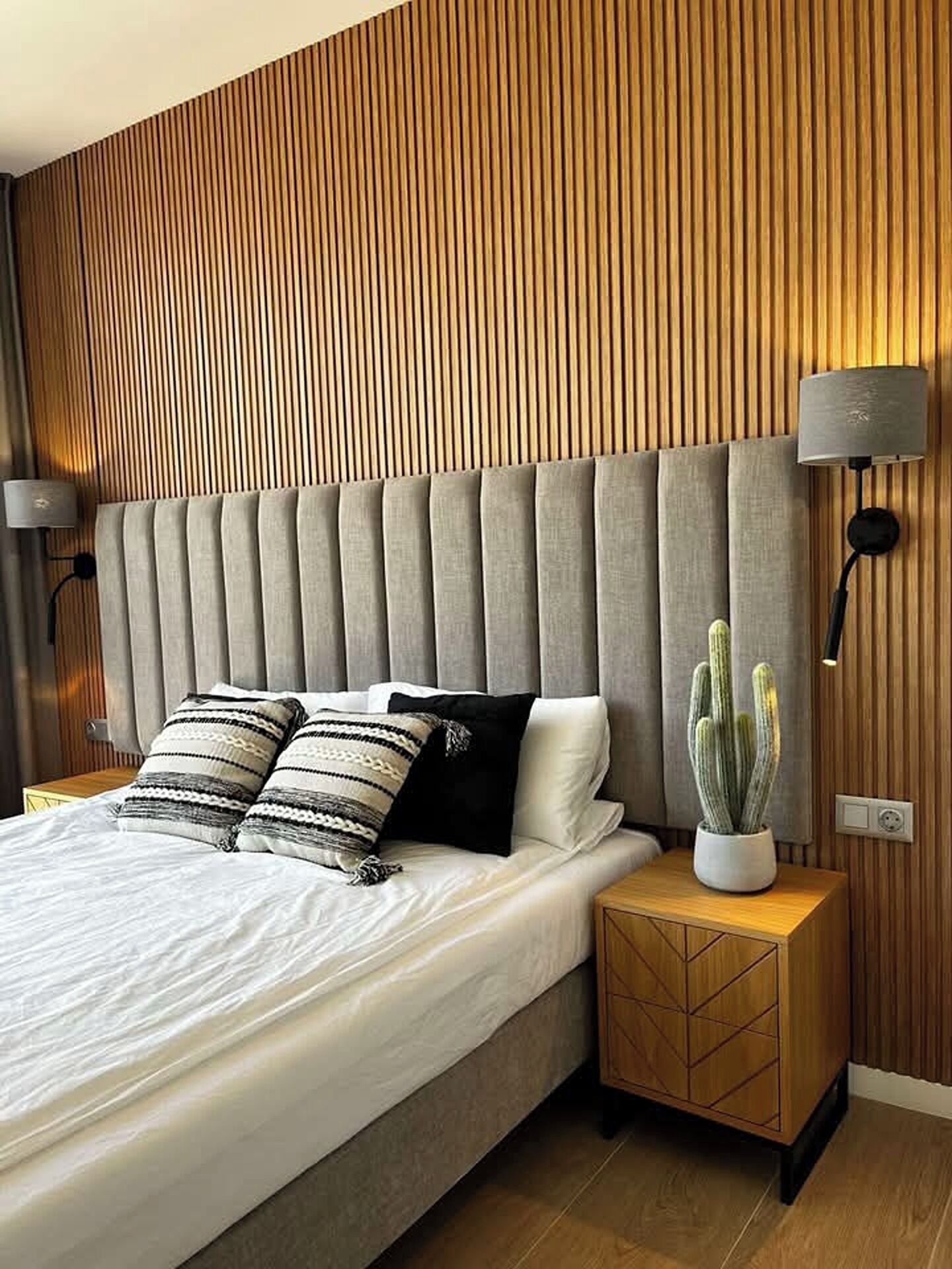 Una camera da letto moderna con un letto grande, una testiera in legno, un comodino con un cactus e lampade a parete.